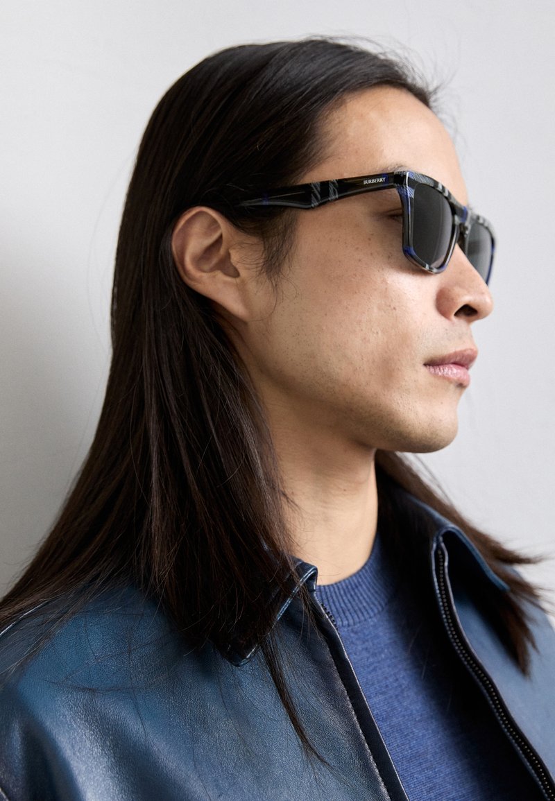 Black and blue patterned sunglasses with dark lenses, worn with a blue sweater and a blue leather jacket, highlighting angular design and texture.