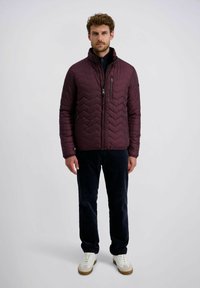 Man in een bordeauxrode gewatteerde jas, zwarte broek en witte sneakers die tegen een effen lichte achtergrond staat.