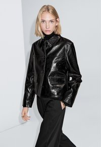 Zwarte jas van nep leer met een doorgestikte voorkant met knopen, een gestructureerde kraag, twee voorzakken en een glanzende afwerking, gecombineerd met een zwarte broek.