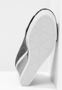 Svart sneakers visad från sidan, vilande mot en vit yta, med en vit sula som har upprepande "Calvin Klein"-logotyp.