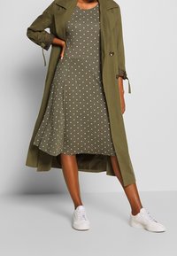 Oliwkowy trencz na zielonej sukience w kropki z rozkloszowanym dołem, zestawiony z białymi trampkami. Gładki materiał, długość do kolan.