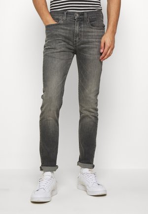 Man wearing faded black slim-fit jeans, striped shirt tucked in, white sneakers, with one hand in pocket and cuffed hems.