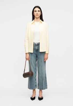 Femme debout portant une chemise boutonnée jaune pâle sur un haut blanc, un jean à jambes larges, des talons noirs, et tenant un sac à main cylindrique marron.