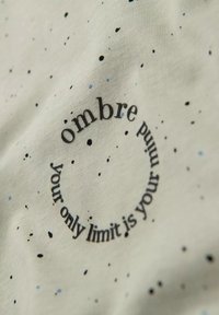 Tejido gris claro con patrón de manchas negras y azules. Presenta un texto circular: "sombre tu único límite es tu mente". Textura suave.
