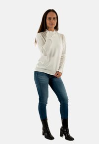 Top bianco a maniche lunghe con perline testurizzate e scollatura arricciata; abbinato a jeans blu e stivali neri con tacco. Design semplice con dettagli sottili.