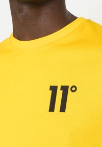 T-shirt in cotone giallo con collo rotondo e logo stampato nero "11°" sul petto. Texture liscia, design casual.