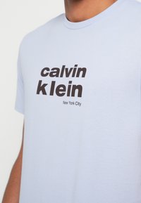 Hele sinine puuvillane t-särk, millel on julged mustad kirjad "calvin klein" ja allpool "New York City", ümmargune kaelus ning lühikesed varrukad.