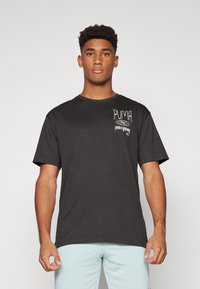 Camiseta negra de manga corta en algodón con corte holgado. Presenta un diseño del logo de Puma en blanco y amarillo en la parte izquierda del pecho.