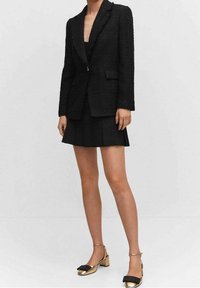 Schwarzer strukturiert Blazer mit Revers und einem Knopf, kombiniert mit einem gefälteten schwarzen Rock und goldenen Blockabsatzschuhen mit einer schwarzen Schleife.