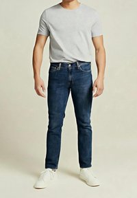 Homme portant un t-shirt gris clair, un jean bleu foncé et des baskets blanches, debout devant un fond uni clair.