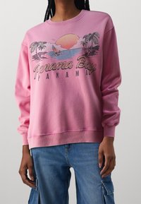 Pink sweatshirt with a round neck and long sleeves, featuring a graphic design of palm trees and a sunset, labeled "Panama Bay."
