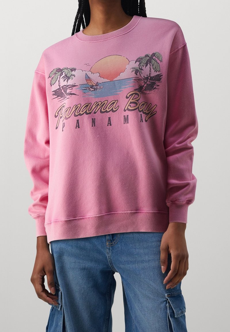 Pink sweatshirt with a round neck and long sleeves, featuring a graphic design of palm trees and a sunset, labeled "Panama Bay."