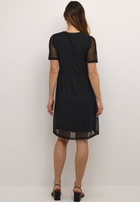 Crna haljina s kratkim prozirnim rukavima, uskim tijelom i lepršavom suknjom. Prozirna tkanina na rubu dodaje detalj slojevitosti.