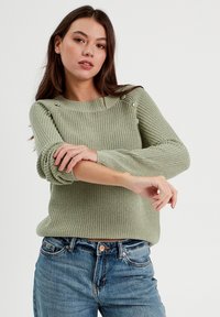 Pull en tricot vert avec une texture côtelée, col rond et accents de boutons sur l'épaule. Accompagné d'un jean en denim bleu.