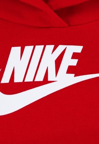Rdeča jopa s kapuco z izrazitim belim napisom "NIKE" in logotipom v obliki kljuke, izdelana iz mehkega blaga z teksturirano površino.