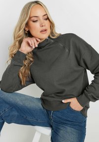 Mörkgrå sweatshirt med hög krage och dragkedjedetalj, tillsammans med blå denimjeans. Har ribbade ärmslut och en avslappnad passform.