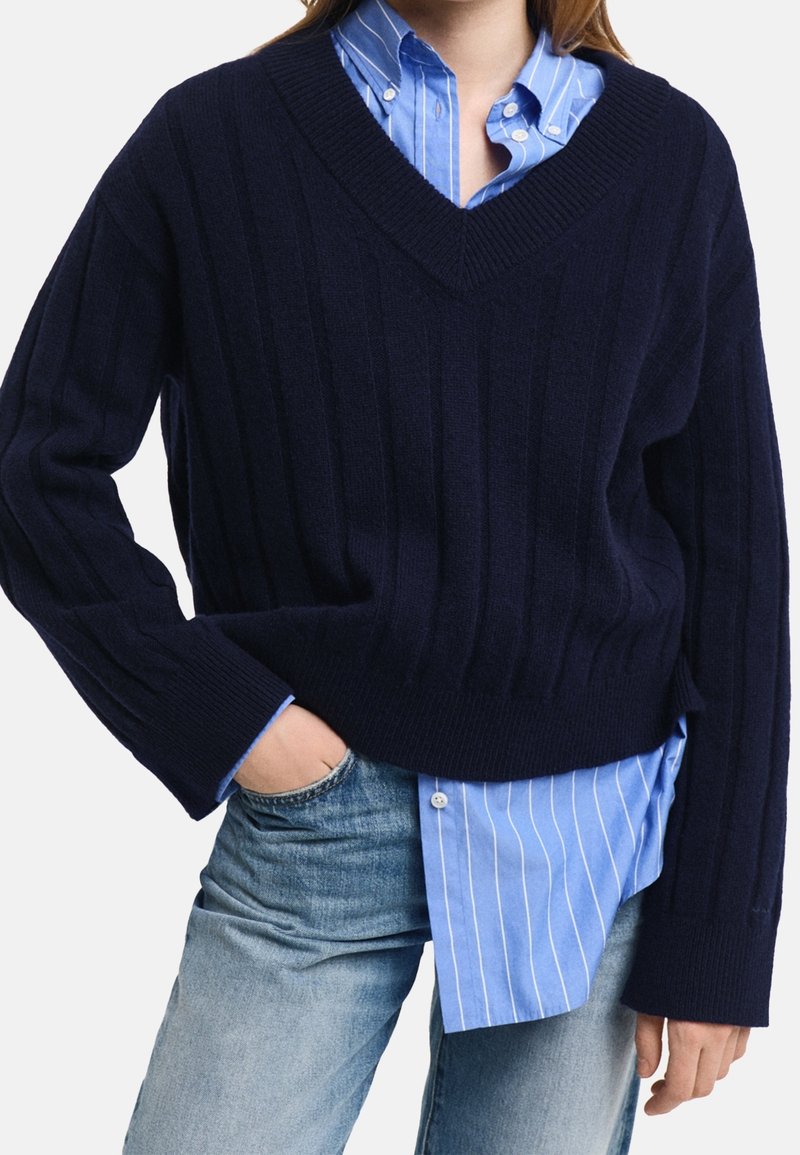 Personne portant un pull côtelé à col en V bleu marine par-dessus une chemise à rayures bleues et blanches avec un jean bleu délavé, une main dans la poche.