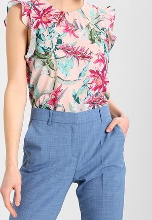 Mujer con blusa sin mangas con estampado floral y volantes en los hombros, y pantalones de tiro alto color azul claro, a medida, con una mano en el bolsillo.