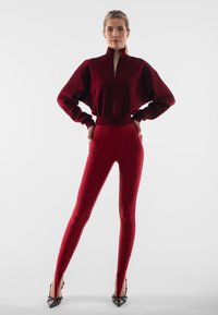 Bordowy skórzany pulower z wysokim kołnierzem i zamkiem błyskawicznym, połączony z dopasowanymi bordowymi legginsami i czarnymi szpilkami z ostrym noskiem. Gładka tekstura.