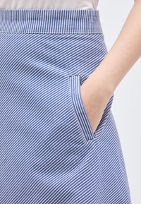 Hand in de zak van een blauw-witte, verticaal gestreepte rok, gecombineerd met een witte top, close-up van stof en stiksels.