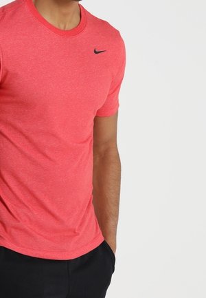 Homme portant un t-shirt rouge Nike avec un pantalon noir, debout avec une main dans sa poche sur un fond clair uni.
