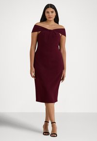 Μπουρντό φόρεμα με έξω τους ώμους και λεπτομέρεια φιόγκου στη λαιμόκοψη, με εφαρμοστή σιλουέτα που στενεύει μέχρι το γόνατο. Φορεμένο με μαύρες ψηλοτάκουνες σανδάλες με λουράκια.