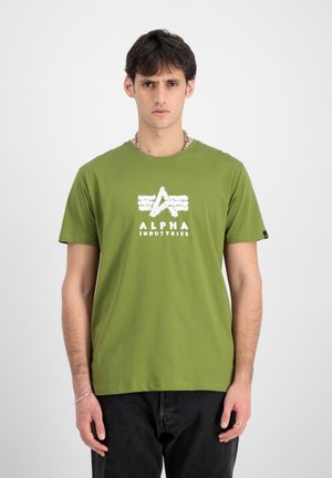 Junger Mann, der ein grünes Alpha Industries T-Shirt, schwarze Hosen, eine silberne Halskette und Ringe trägt, vor einem schlichten weißen Hintergrund steht.