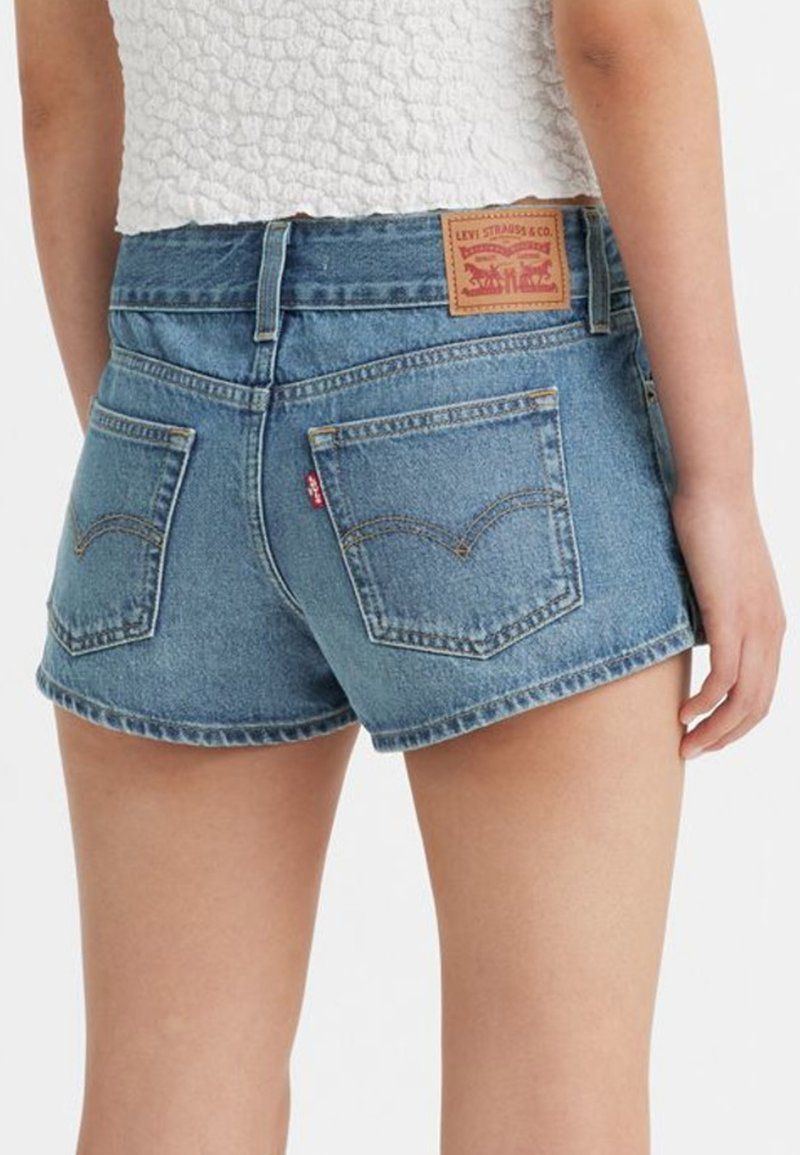 Personne portant un short en denim bleu Levi's et un haut blanc texturé, montré de la taille vers le bas, vue de dos sur fond blanc.