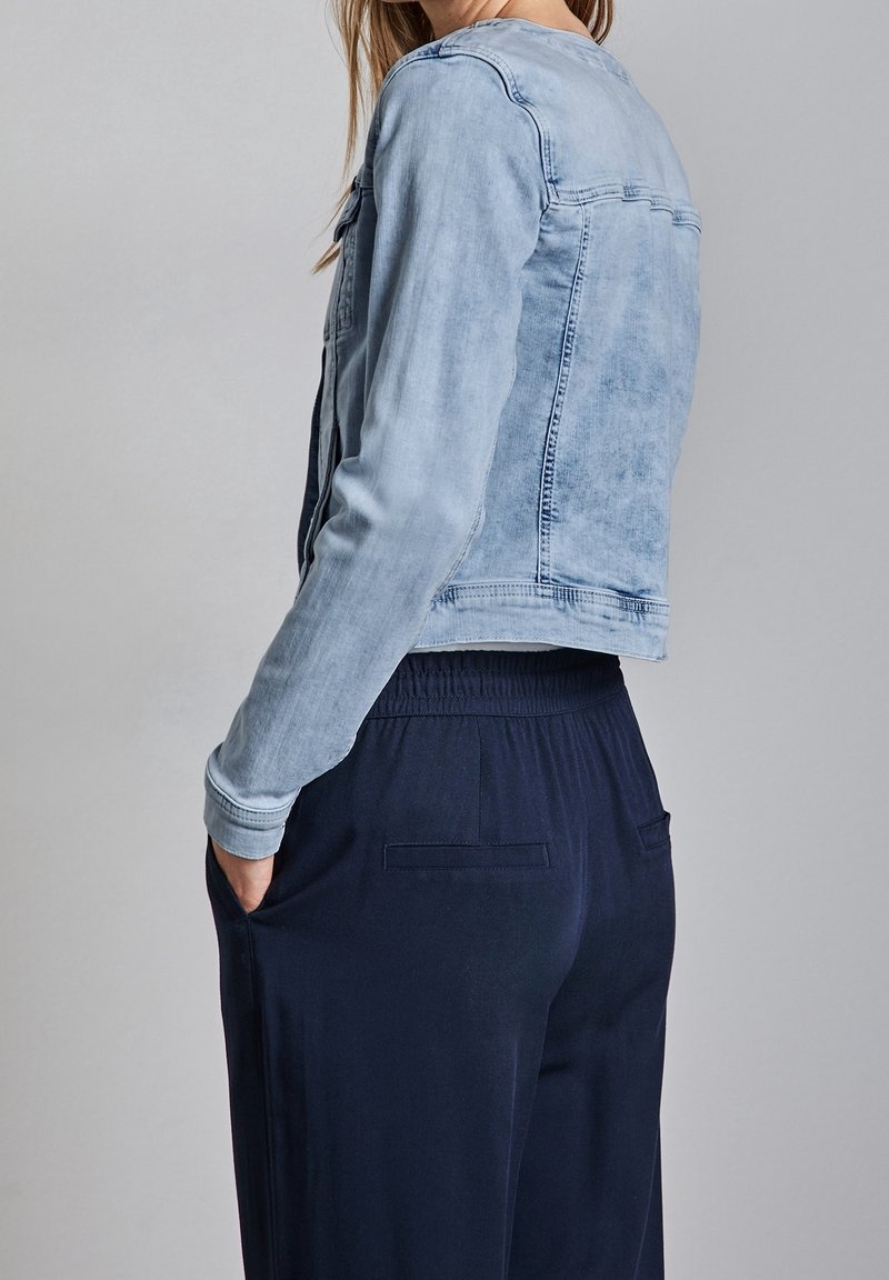 Person wearing a light blue denim jacket with long sleeves and dark navy pants with hands in side pockets against a plain background.