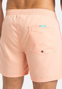 Shorts de bain couleur pêche avec une taille élastique, dotés d'une poche arrière, d'un matériel en métal circulaire et d'une texture de tissu lisse.