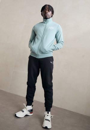 Veste zippée vert menthe avec col montant, pantalon de survêtement noir et baskets blanches avec accents noirs. Logo Puma présent sur tous les articles.