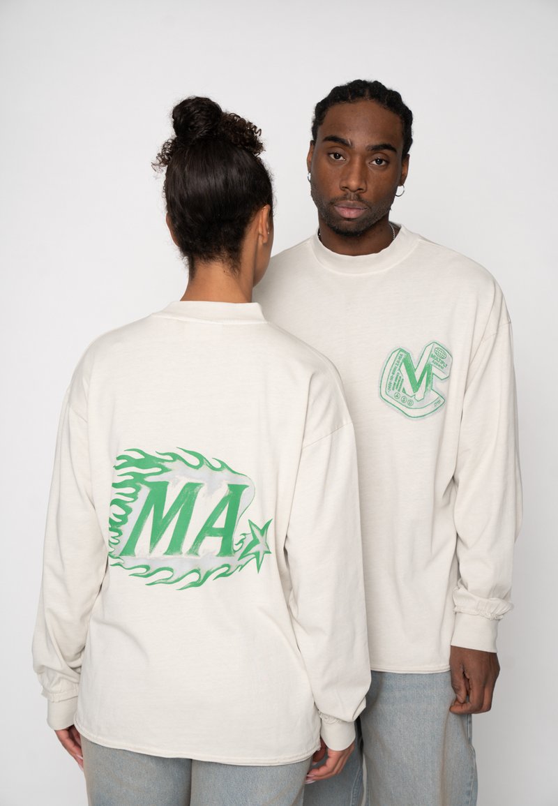 Long-sleeve beige t-shirts featuring green graphics; one displays "MA" with a flame design, the other has a decorative letter "C."