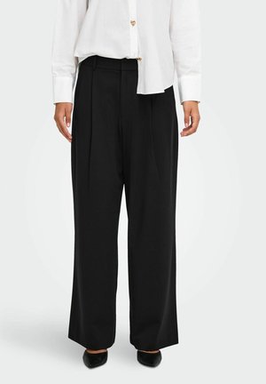 Femme portant un pantalon noir taille haute à jambes larges, associé à un chemisier blanc boutonné et des chaussures noires pointues.