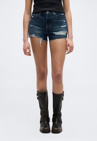 Γυναίκα που φοράει σκισμένα σκούρα μπλε τζιν σορτς, μαύρο αμάνικο τοπ και μαύρες δερμάτινες μπότες μέχρι το γόνατο, στέκεται μπροστά σε λευκό φόντο.