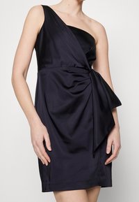 Jednokrčný tmavě fialové saténové šaty s přiléhavým živůtkem, s drapovaným detailem látky a zavazovacím akcentem v pase. Hladká textura.