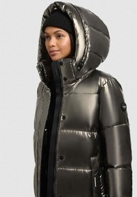 Vrouw in een glanzende donkere metallische pufferjack met een grote capuchon en een zwarte gebreide muts, die naar links kijkt tegen een effen achtergrond.
