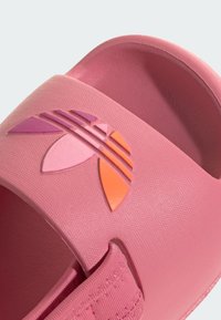 Vaaleanpunaiset liukuvarpaat, joissa on teksturoitu pinta. Ominaisuutena selkeästi painettu logo purppuran ja oranssin sävyissä. Pehmeä, joustava hihnasuunnittelu.