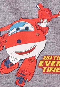 Grafica di un aereo cartoon rosso e bianco su tessuto grigio. Caratterizzato da occhi grandi, bocca sorridente e testo "IN TEMPO OGNI VOLTA" in giallo in grassetto.