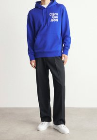 Koninklijk blauwe hoodie met het witte "Calvin Klein Jeans" logo, buidelzak, gedragen met zwarte broek en witte sneakers.