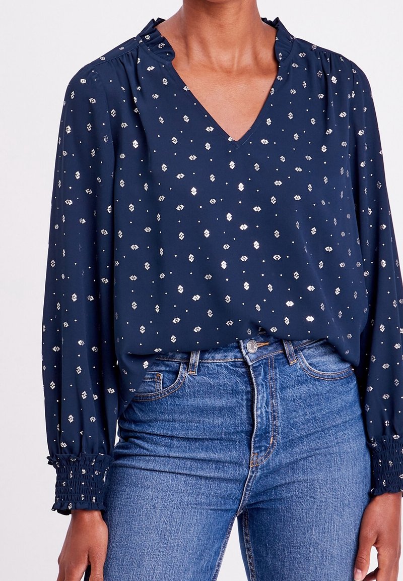Femme portant un chemisier bleu marine à manches longues avec motif losanges blancs, poignets smockés, col à volants, rentrée dans un jean bleu taille haute.