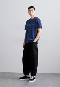 Marineblå bomull t-skjorte med "HELMUT LANG" trykket i grått, kombinert med svarte vide bukser og svarte joggesko med hvite såler.