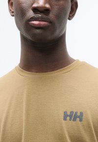 T-shirt à manches courtes de couleur brun clair avec un col rond. Présente un logo gris imprimé sur la partie gauche de la poitrine. Texture douce et lisse.