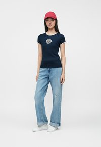 Γυναίκα που φοράει ένα κόκκινο καπέλο, ναυτικό μπλε κοντομάνικο πουκάμισο, ανοιχτό μπλε τζιν με καμπάνι και λευκά αθλητικά παπούτσια, στέκεται μπροστά σε ένα απλό φόντο.