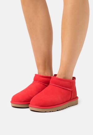 Jambes portant des bottines rouges en suède montant à la cheville, avec des semelles en caoutchouc beige, posées sur un fond uni gris clair.