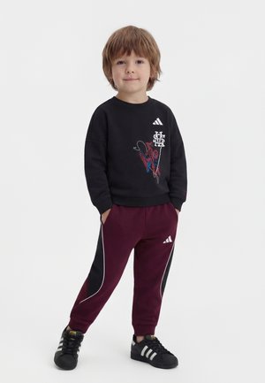 Młody chłopiec stojący z rękami w kieszeniach, ubrany w czarną bluzę Spider-Man, bordowe spodnie dresowe Adidas i czarne trampki Adidas.