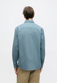 Srajca z dolgimi rokavi iz mehkega, svetlo modrega bombaža s klasičnim ovratnikom, gumbi na manšetah in ukrivljenim robom, prikazana s hrbta.