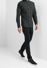 Muž kráčející v tmavě zelené kostkované košili na knoflíky, černých kalhotách a černých teniskách s bílými podrážkami na jednobarevném pozadí.