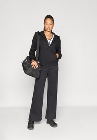 Μαύρο φούτερ με φερμουάρ και παντελόνι σε φαρδιά γραμμή από απαλό ύφασμα, σετ με μαύρη υφασμάτινη τσάντα ώμου και μαύρα αθλητικά παπούτσια.