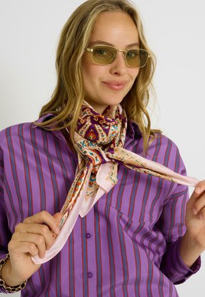 Mujer con gafas de cristales tintados en amarillo, camisa con rayas moradas y pañuelo floral multicolor anudado alrededor del cuello, sujetando los extremos del pañuelo con ambas manos.