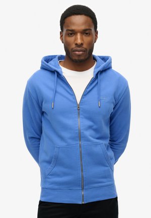 Sudadera azul con cremallera y capucha ajustable, bolsillos frontales y puños acanalados. Textura suave con un sutil logo bordado en el pecho.